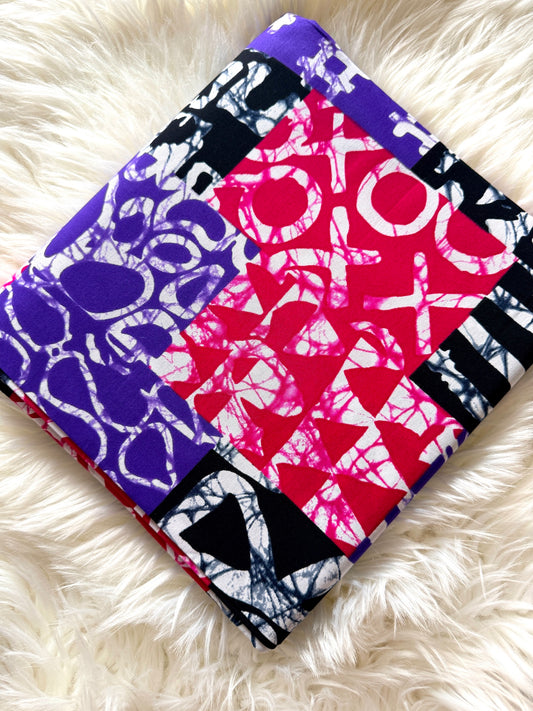 A square fabric item, likely a small quilt, blanket, or pillow cover, featuring a bold patchwork design of red, purple, black, and white patterns, displayed on a white faux fur surface.