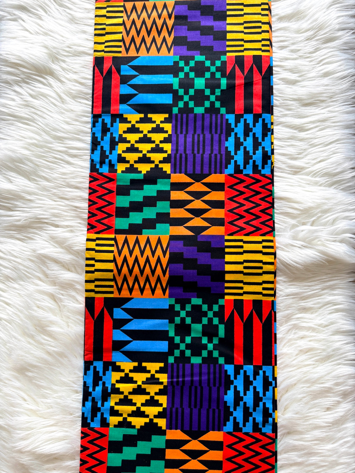 A long rectangular textile laid flat on a white faux-fur surface, featuring a bold patchwork of geometric and zigzag patterns in yellow, black, red, blue, purple, and teal.