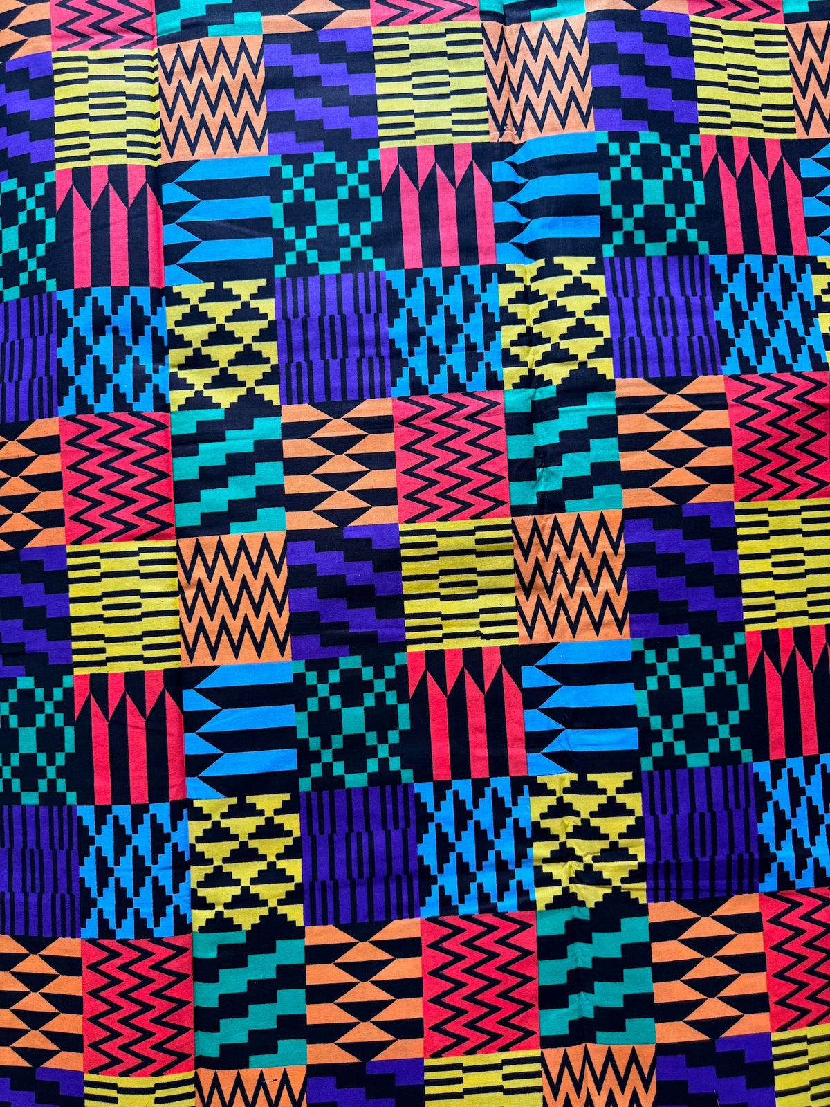 A close-up of a vibrant, multicolored geometric fabric featuring a patchwork of African Kente patterns in pink, yellow, blue, purple, orange, and black.