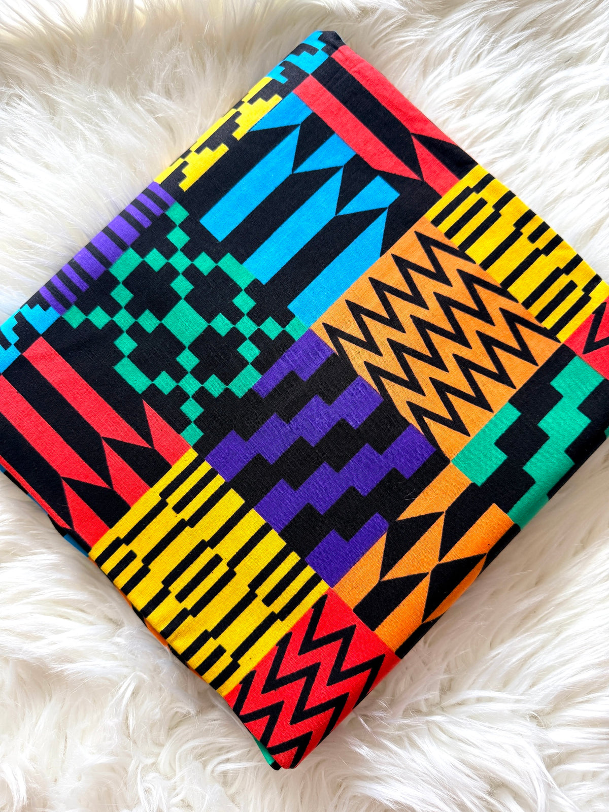 A folded square fabric item, likely a scarf or small blanket, featuring a vibrant multicolor geometric print with black, yellow, red, blue, purple, and green patterns, laid flat on a white faux fur surface.
