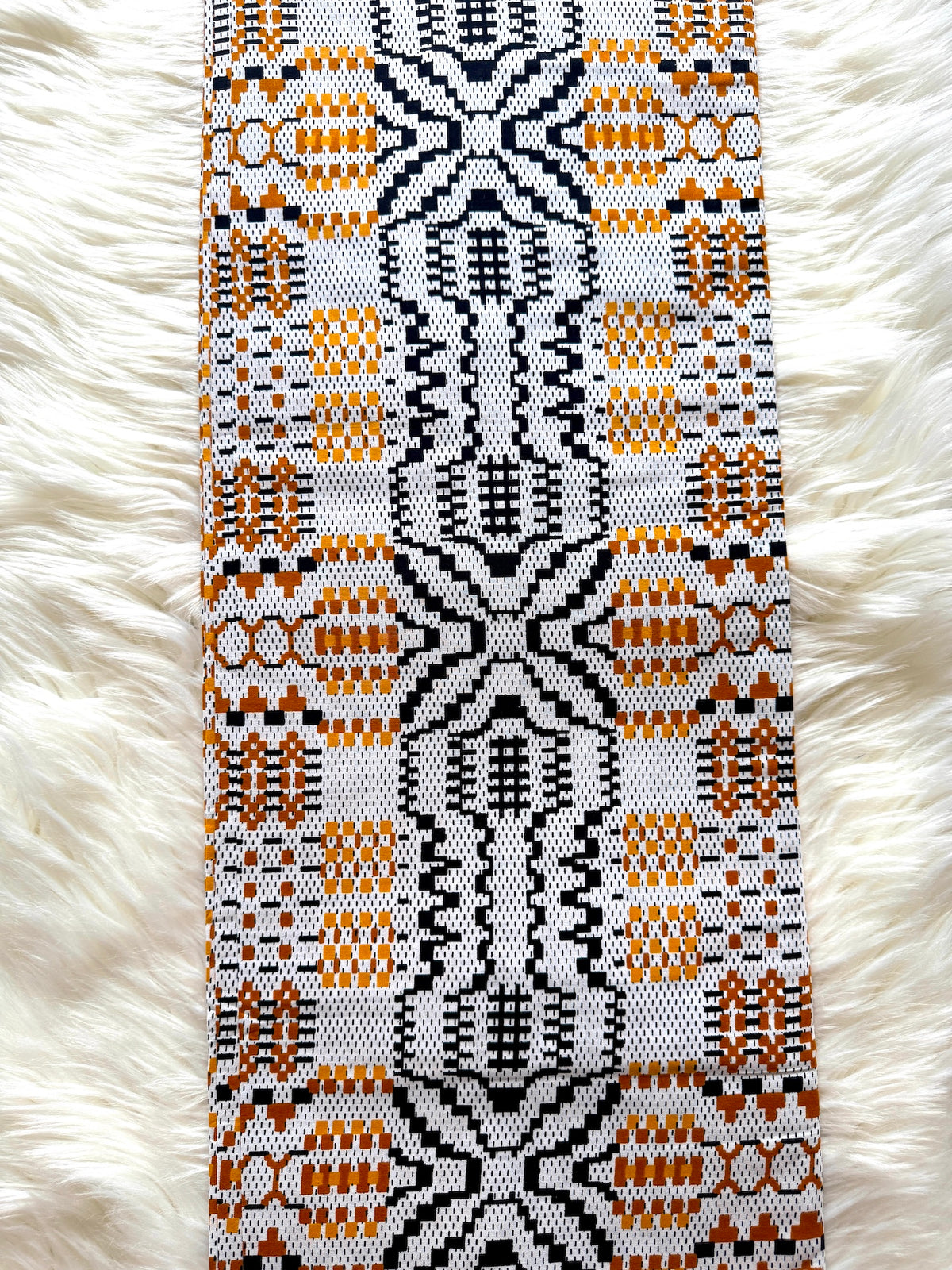 A close-up of a woven textile strip with a geometric pattern in black, orange, and white, laid flat on a white faux fur background.