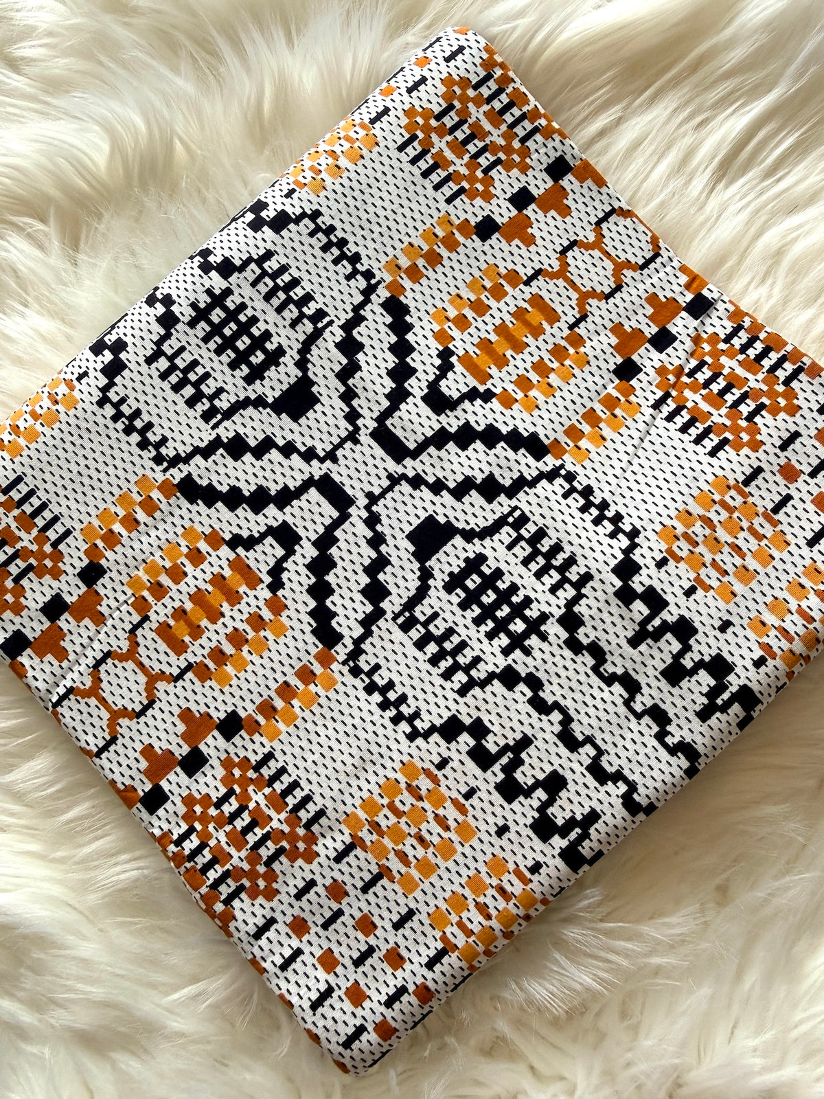 A square woven throw pillow with a geometric pattern in black, orange, and white, displayed on a fluffy white faux fur surface.