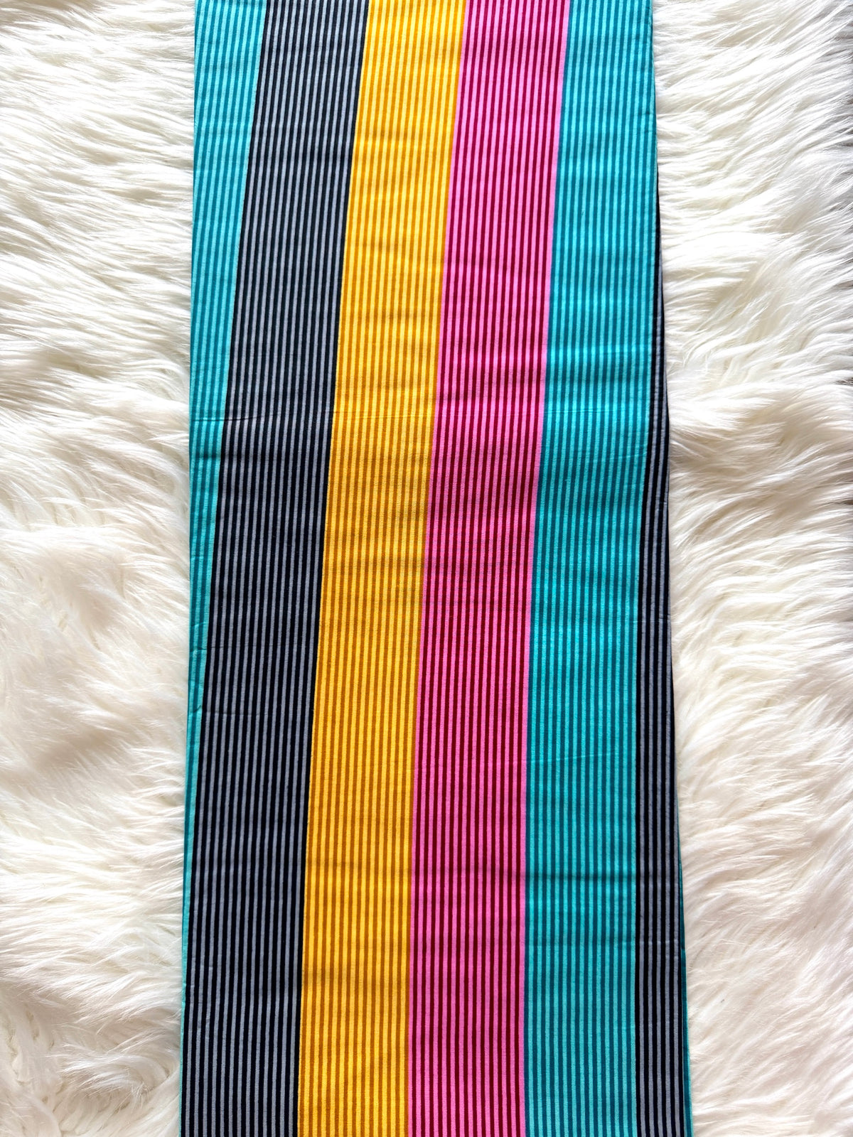 A vertical striped fabric accessory, likely a sash or scarf, featuring alternating stripes of black and white, bright yellow, magenta pink, and turquoise blue, laid flat on a white faux fur background.