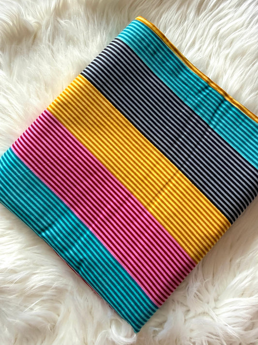 A square fabric item, likely a small pouch or bag, featuring bold horizontal stripes in teal, yellow, pink, and black, laid flat on a white faux fur surface.