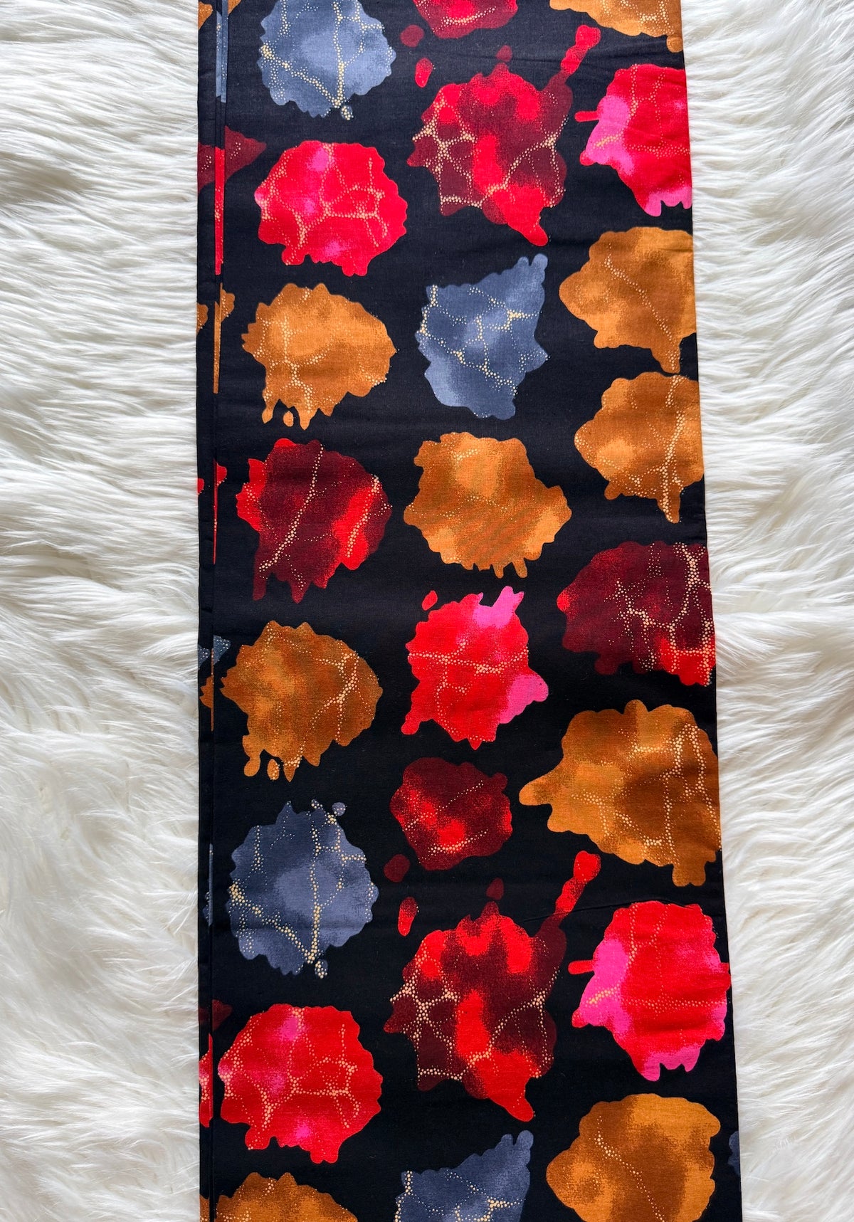 A long, narrow scarf or sash laid flat on a white faux-fur surface. The fabric has a black base with large abstract watercolor-like spots in red, orange, pink, and muted blue-gray.