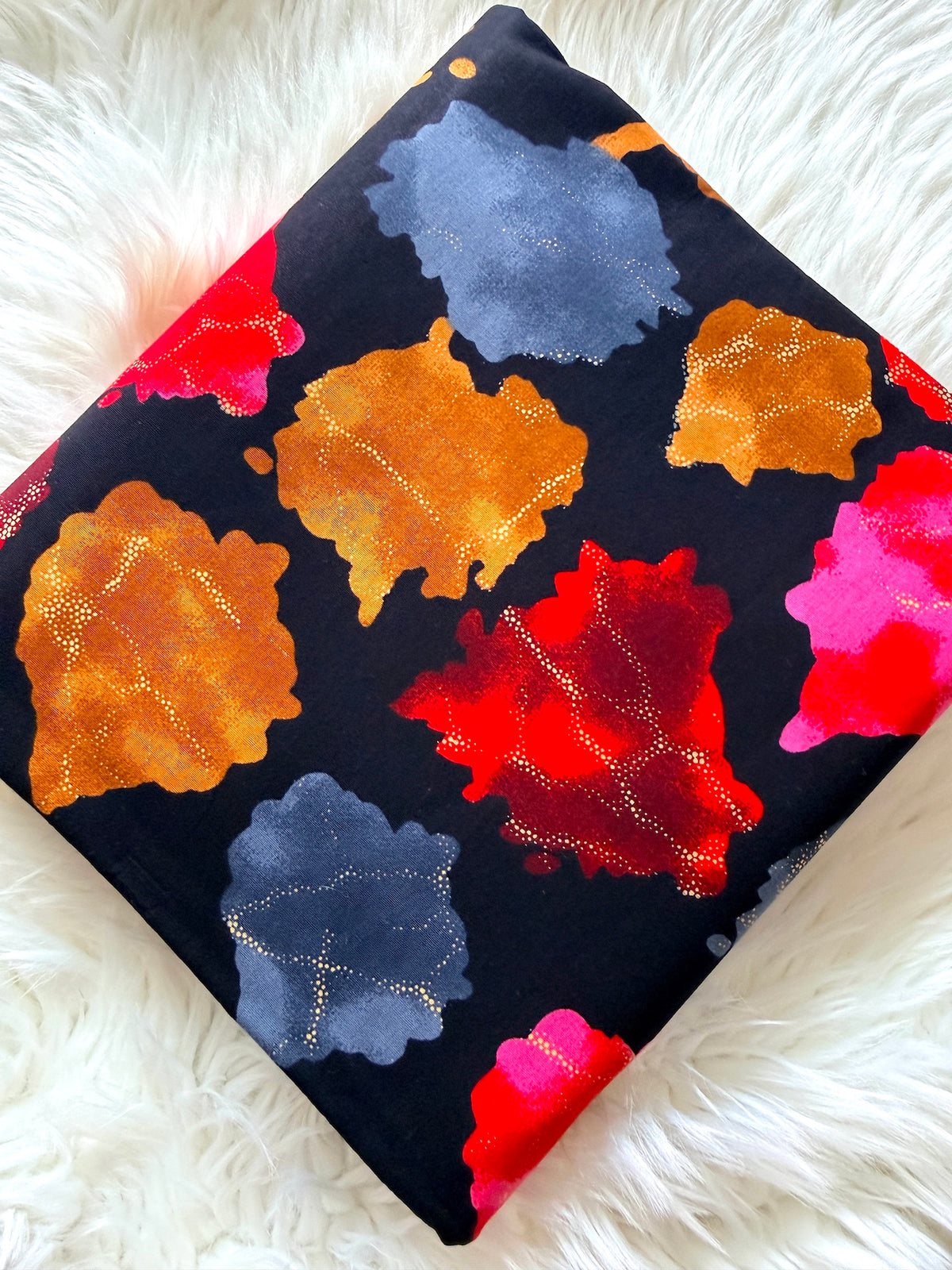 A square throw pillow with a black background and a colorful abstract floral pattern, resting on a white faux fur surface.
