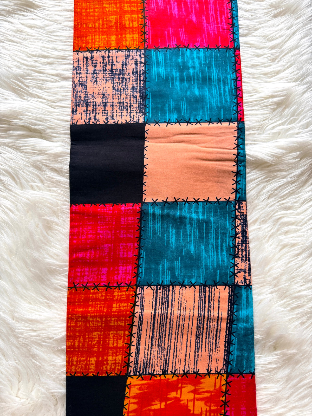 A long rectangular patchwork textile laid flat on a white faux-fur surface. The fabric features multiple square panels in bright orange, pink, turquoise blue, peach, and black, separated by black stitched seams.