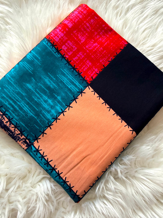 A square patchwork fabric or textile laid flat on a white faux-fur surface, featuring four large color blocks: bright red, teal blue, peach, and black, separated by black stitched borders.