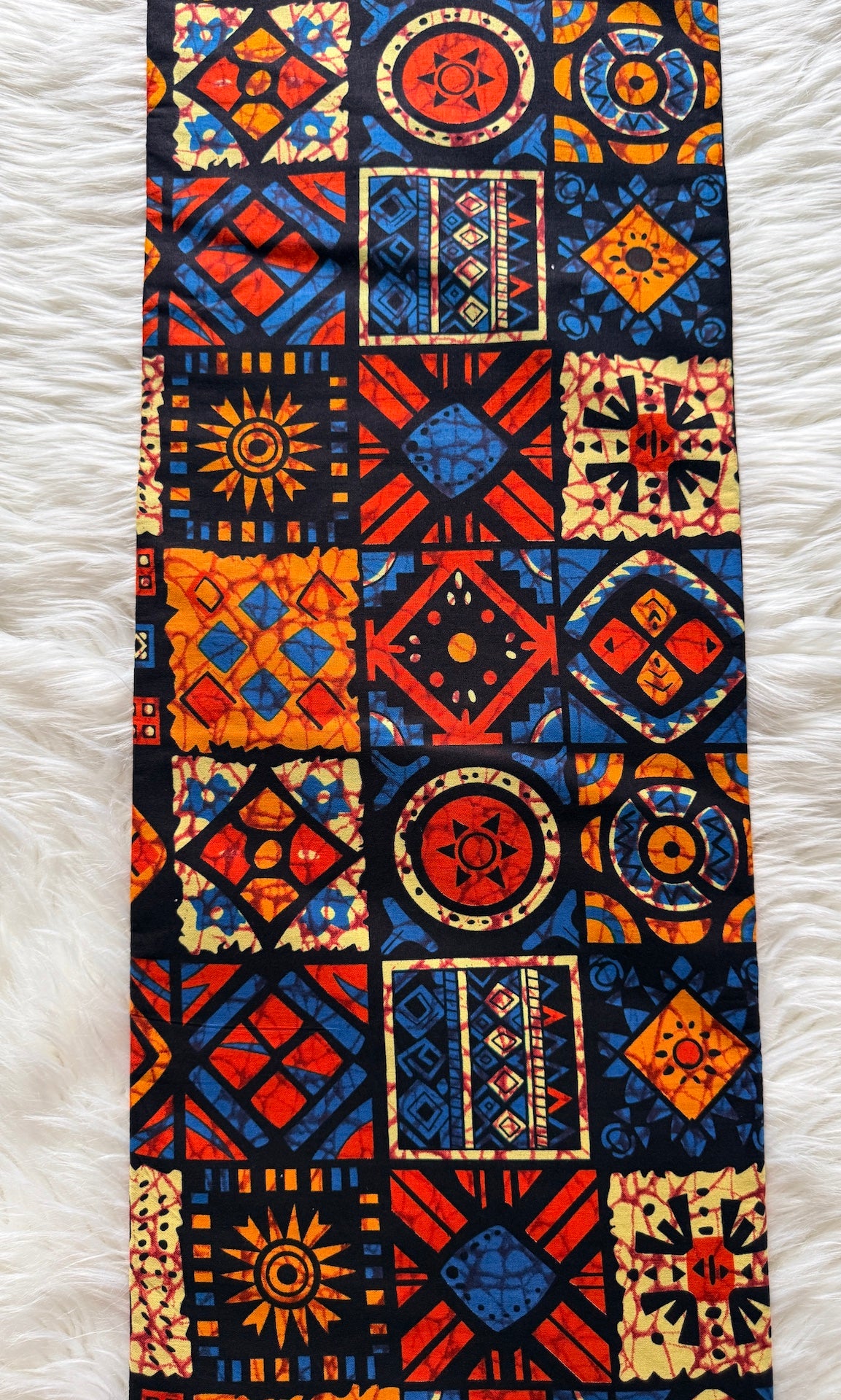 A long rectangular textile with a bold patchwork-style geometric print in red, blue, orange, black, and cream, laid flat on a white faux-fur surface.