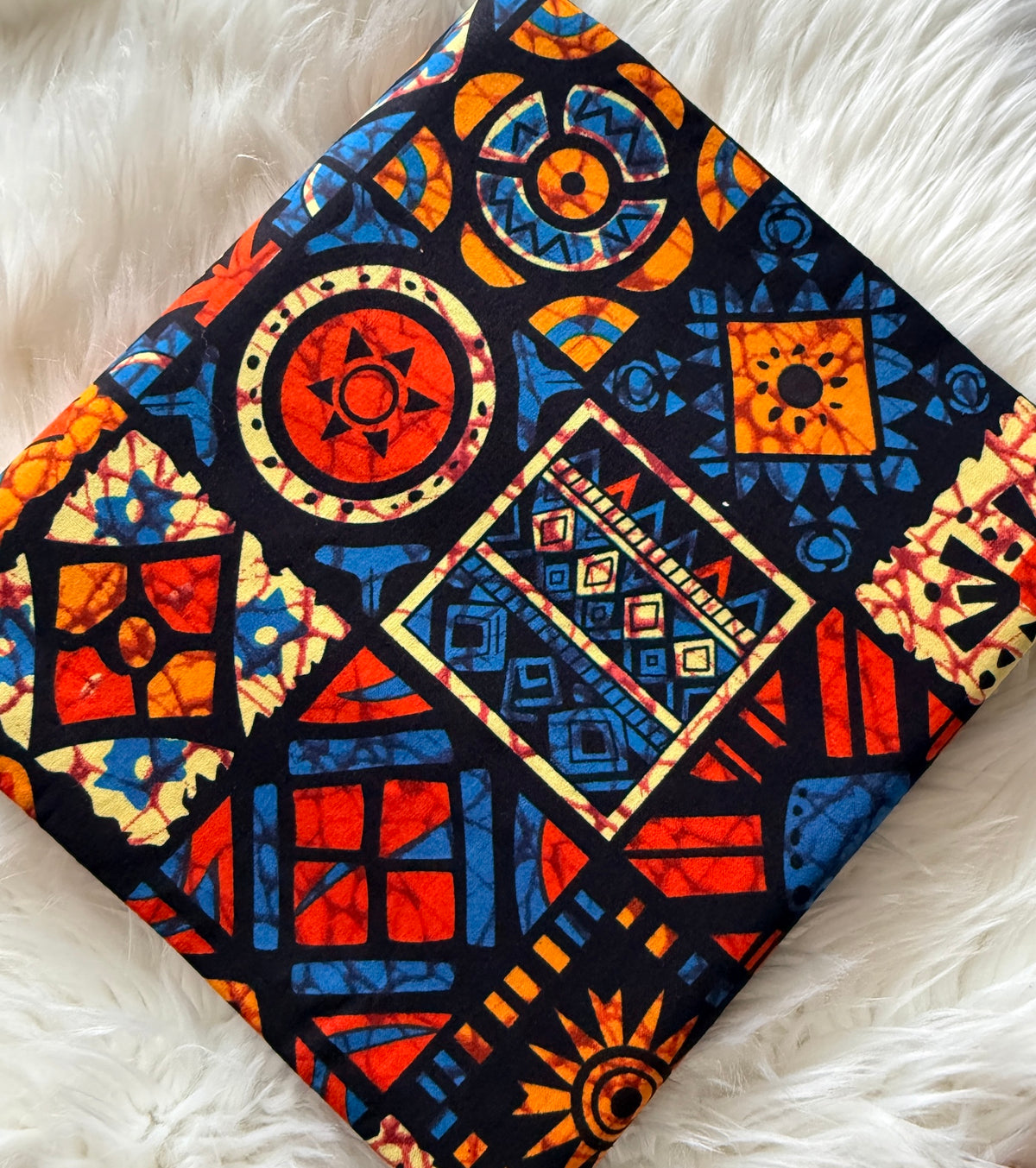 A square fabric item, likely a napkin or small scarf, featuring a vibrant African wax print with geometric and tribal patterns in red, blue, orange, and yellow on a black background, laid flat on a white faux fur surface.