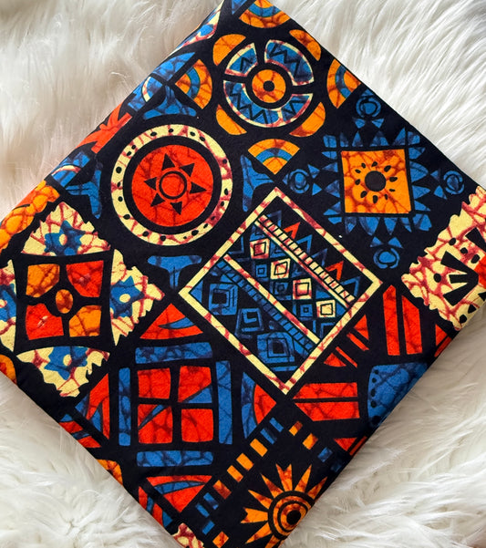 A square fabric item, likely a napkin or small scarf, featuring a vibrant African wax print with geometric and tribal patterns in red, blue, orange, and yellow on a black background, laid flat on a white faux fur surface.
