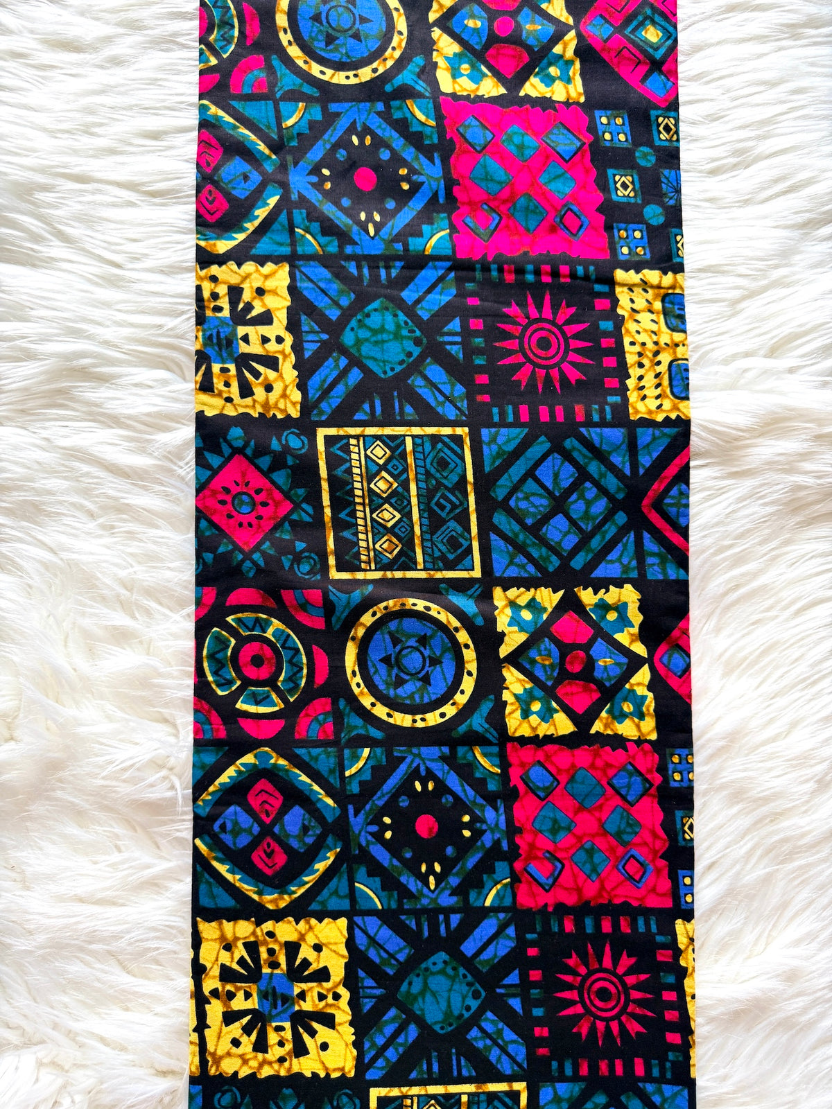 A long rectangular scarf with a vibrant, multicolor geometric patchwork print, laid flat on a white faux fur surface.