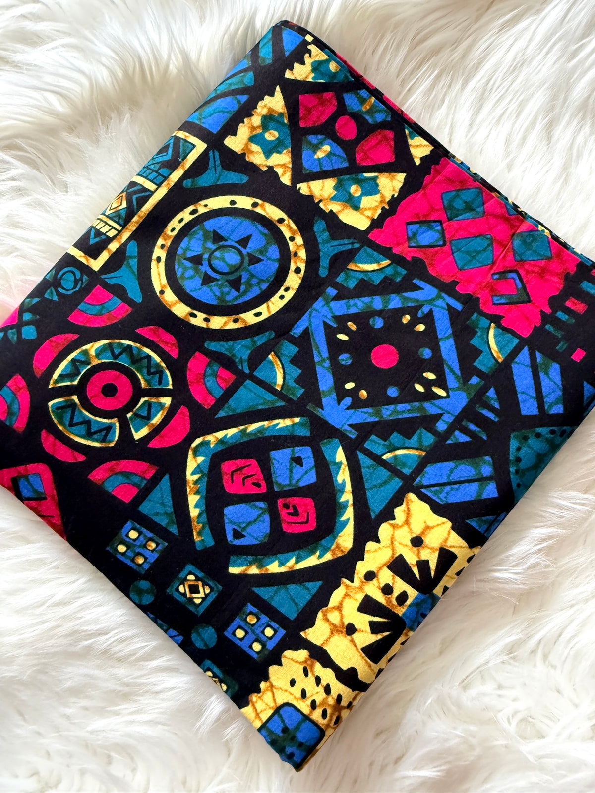 A square fabric item, likely a pillow cover or small quilt, featuring a vibrant, multicolored geometric pattern with circles, diamonds, and triangles in blue, yellow, pink, and red on a black background, laid flat on a white faux fur surface.