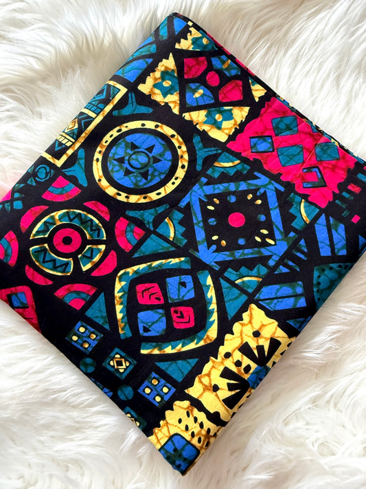 A square fabric item, likely a pillow cover or small quilt, featuring a vibrant, multicolored geometric pattern with circles, diamonds, and triangles in blue, yellow, pink, and red on a black background, laid flat on a white faux fur surface.