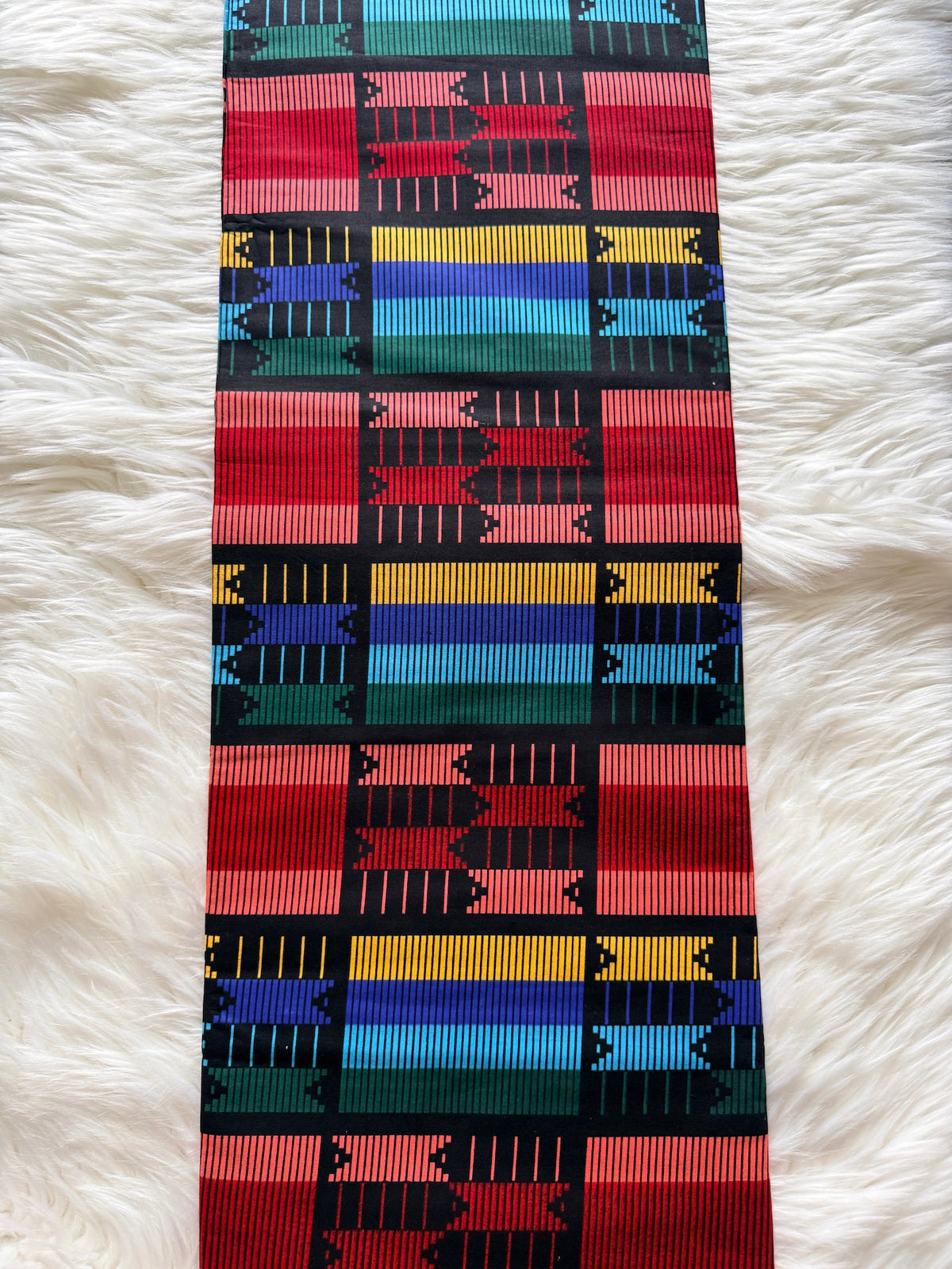 A long woven textile strip with a bold geometric pattern in red, pink, black, blue, yellow, and teal, laid flat on a white faux-fur surface.
