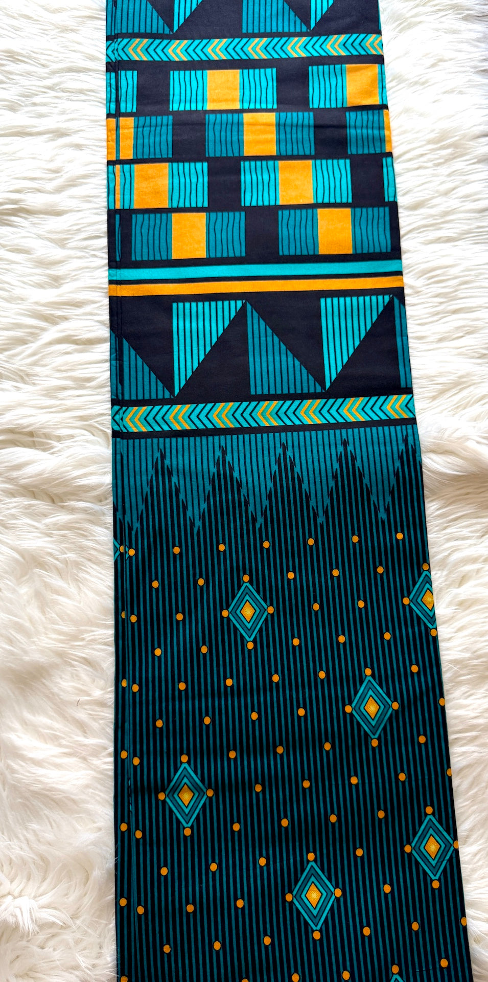 A long fabric accessory, likely a sash or scarf, featuring a vibrant geometric and ikat-inspired pattern in teal, black, yellow, and orange, laid flat on a white faux fur background.