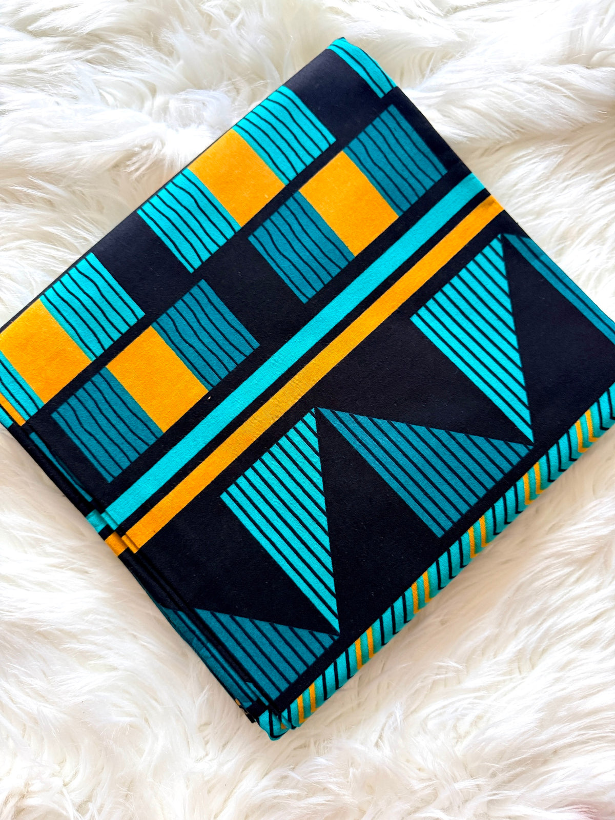 A folded textile with a bold geometric pattern in black, turquoise, and mustard yellow, displayed on a white faux fur surface.