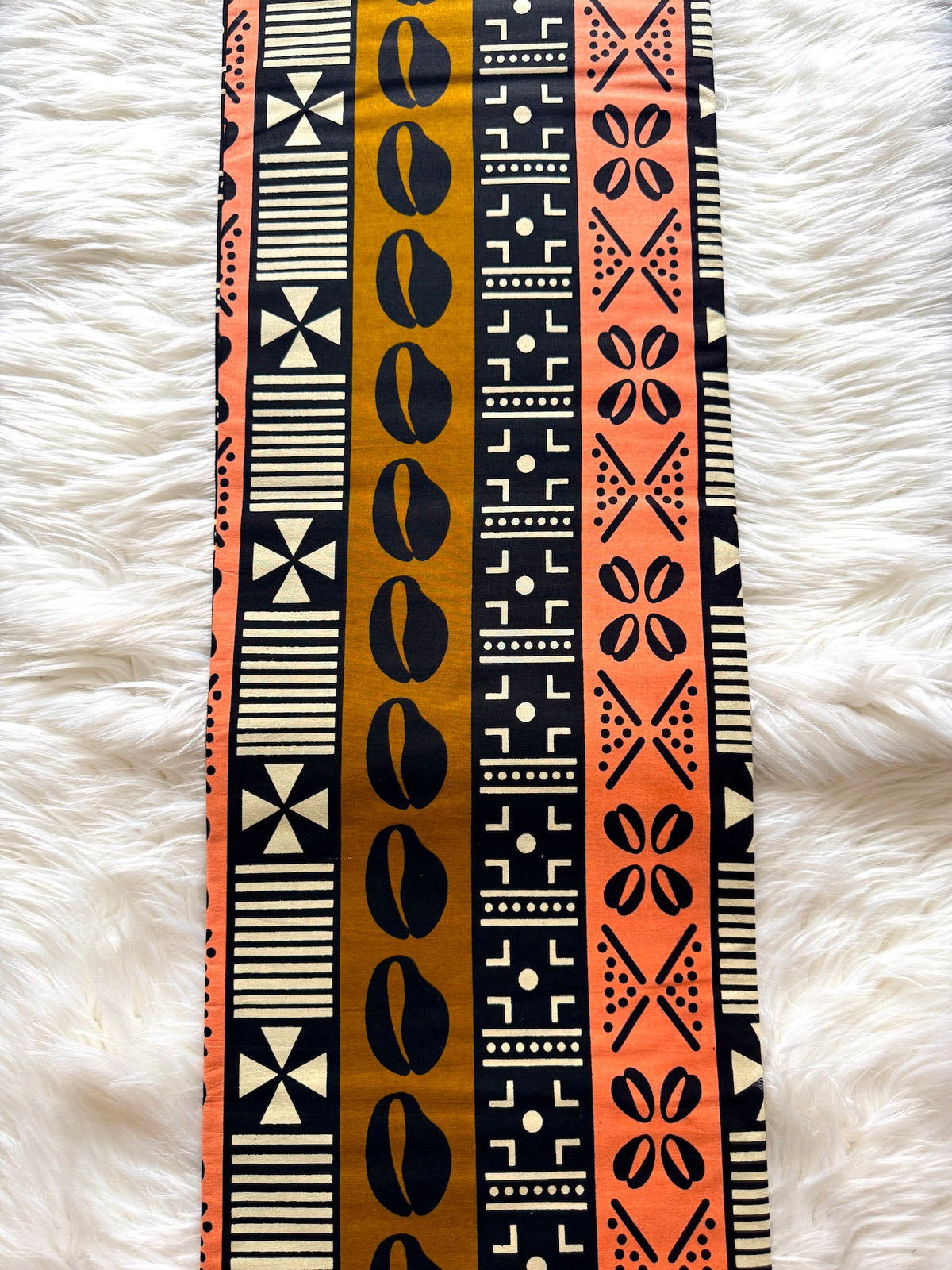 A vertical textile runner or scarf featuring bold vertical stripes with geometric and tribal-inspired patterns in black, orange, beige, and mustard yellow, laid flat on a white faux fur background.