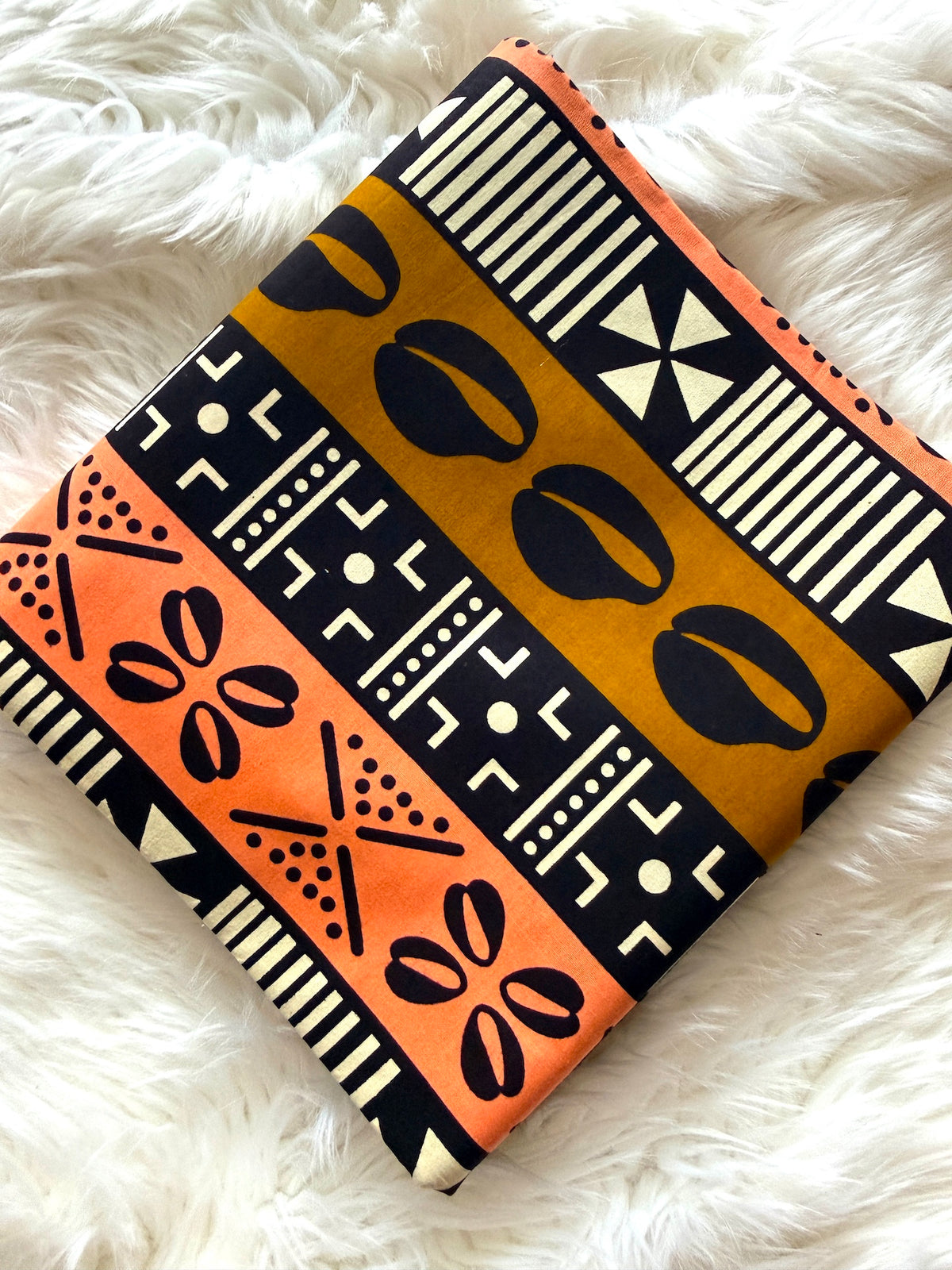 A square fabric pouch or clutch with a bold African-inspired geometric pattern in orange, mustard yellow, black, and cream, resting on a white faux fur surface.