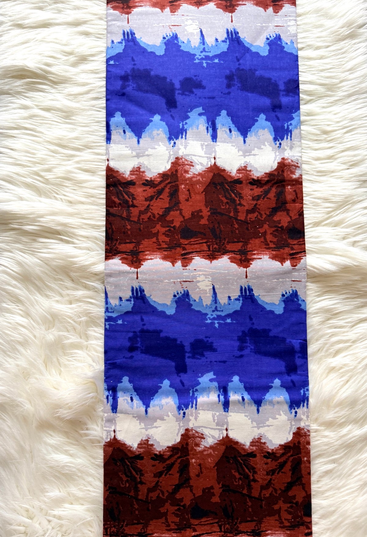 A vertical rectangular textile with an abstract mountain-like pattern in blue, white, and rust red, laid flat on a white faux-fur surface.