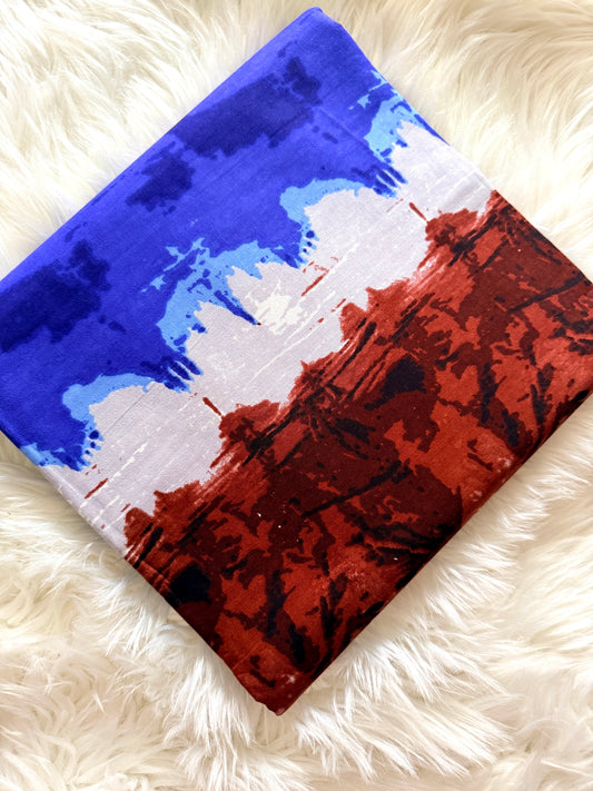 A square fabric item, likely a scarf or small bag, featuring an abstract paint-splatter design in blue, white, and reddish-brown, laid flat on a white faux fur surface.