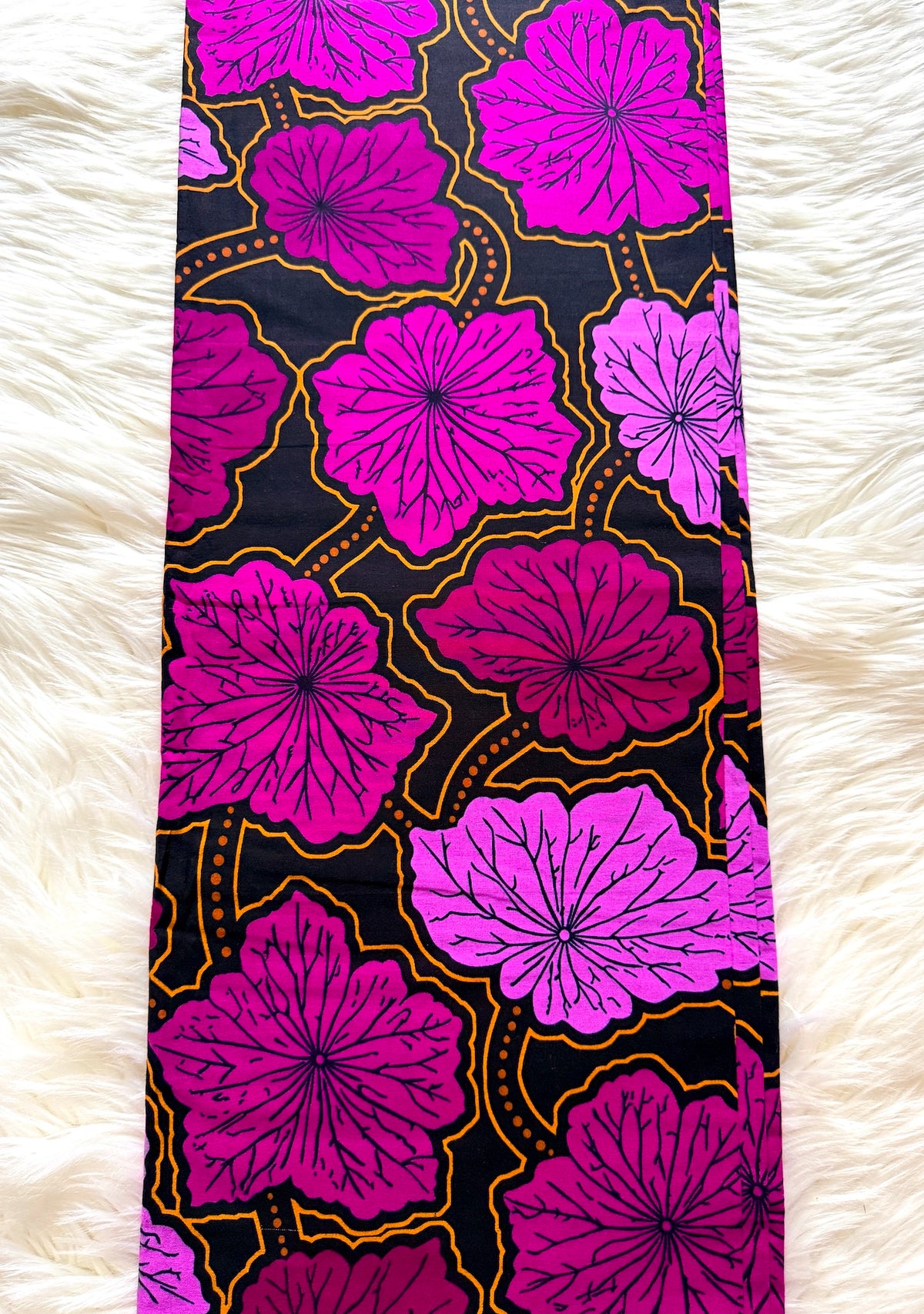 A long rectangular textile laid flat on a white faux-fur surface, featuring a bold floral print with large pink and magenta flowers outlined in yellow on a black background.