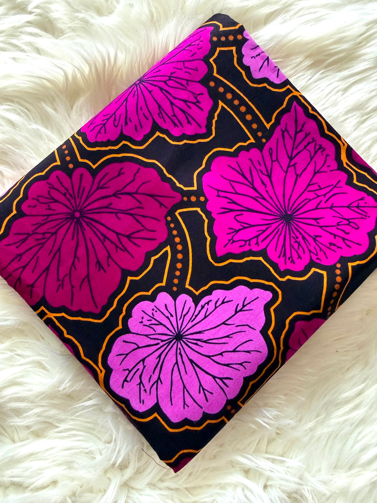 A square piece of dark patterned fabric with large pink and magenta floral motifs outlined in gold, laid flat on a white faux-fur surface.