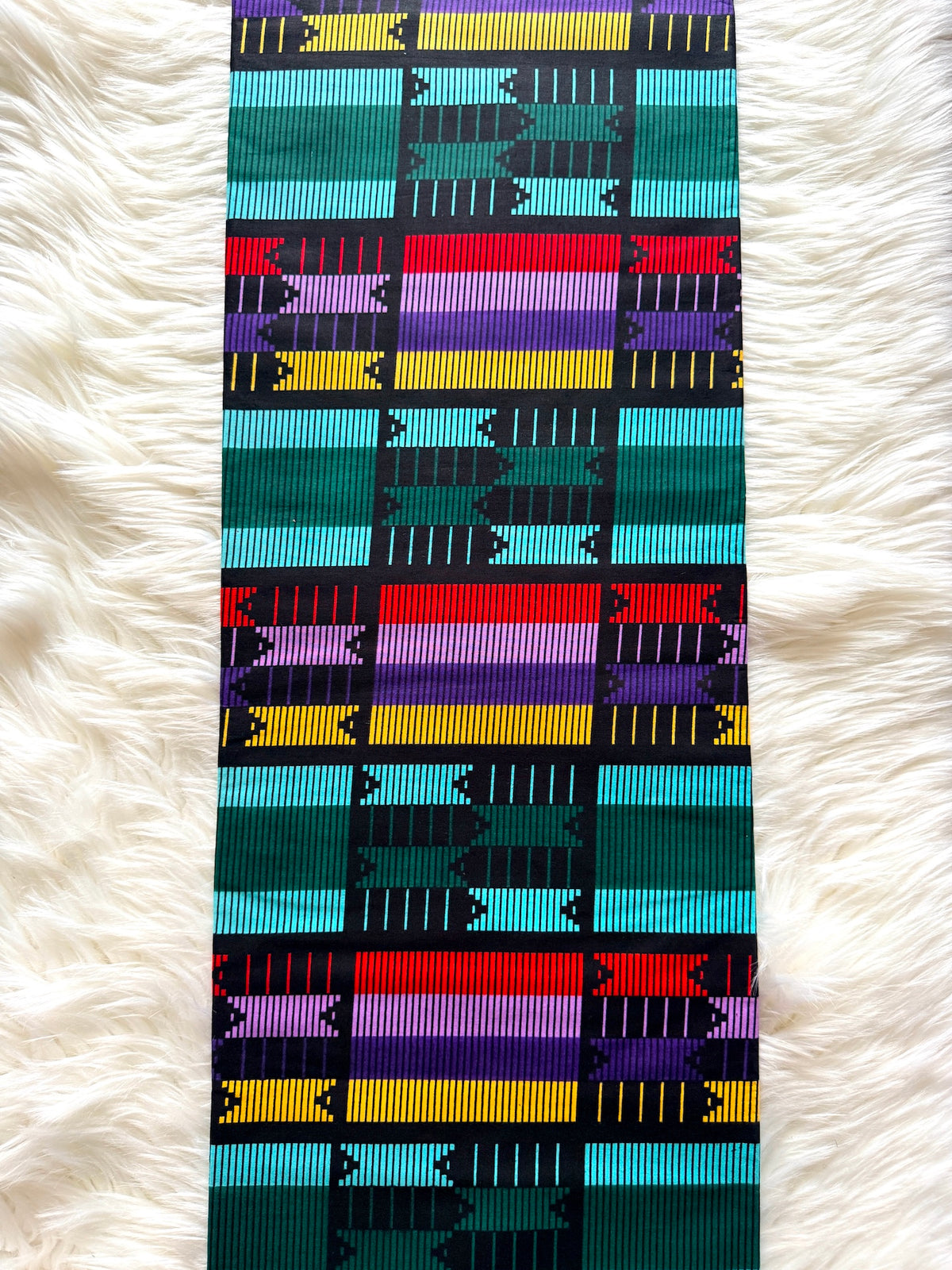 A long rectangular textile with a bold geometric woven pattern in teal, turquoise, black, red, purple, yellow, and white, laid flat on a white faux-fur surface.