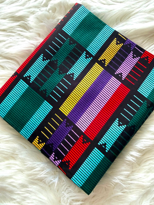 A square woven textile with a bold geometric pattern in teal, black, red, yellow, purple, and white, laid flat on a white faux fur surface.