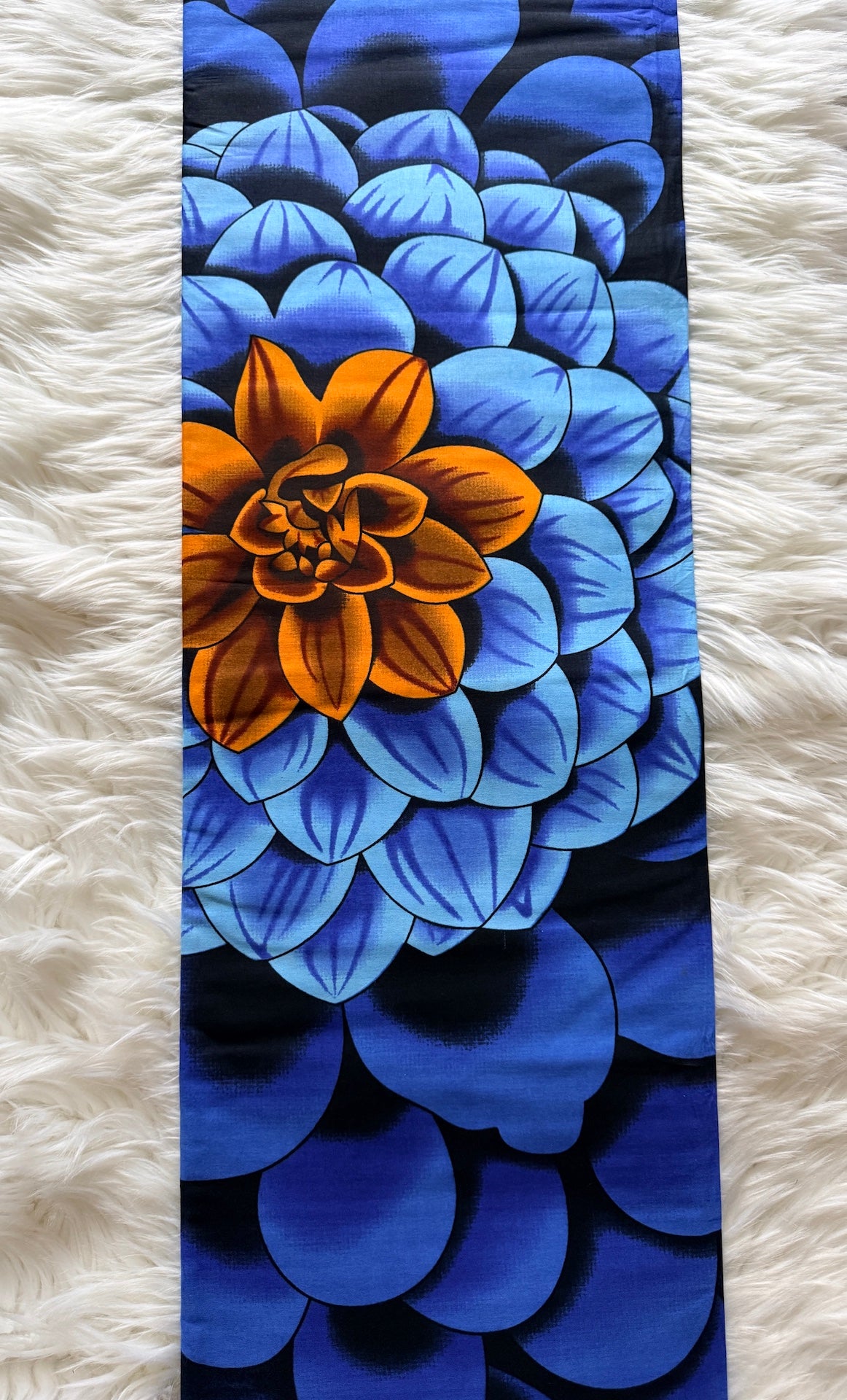 A vertical fabric panel or scarf featuring a bold graphic print of a large orange flower surrounded by layered blue and navy petals, laid flat on a white faux fur surface.