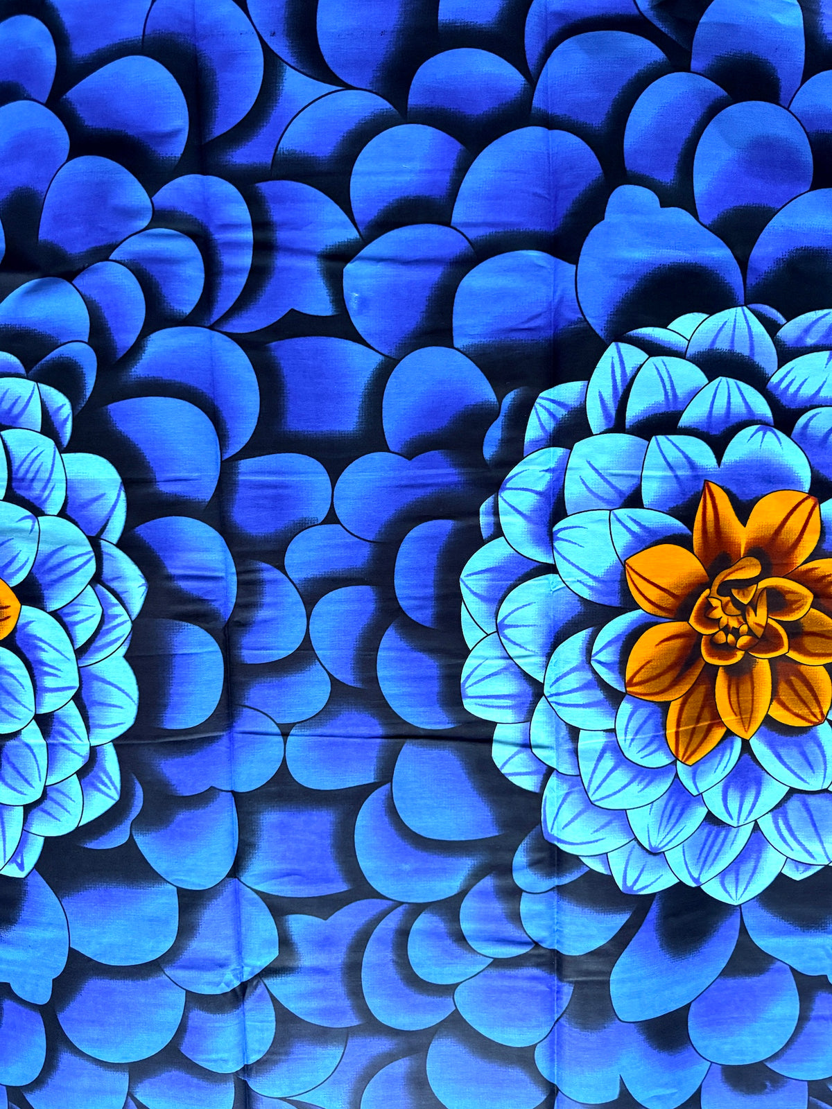 A close-up of a blue patterned fabric or wallpaper featuring a repeating scalloped petal design with two large flower motifs in light blue and bright orange.