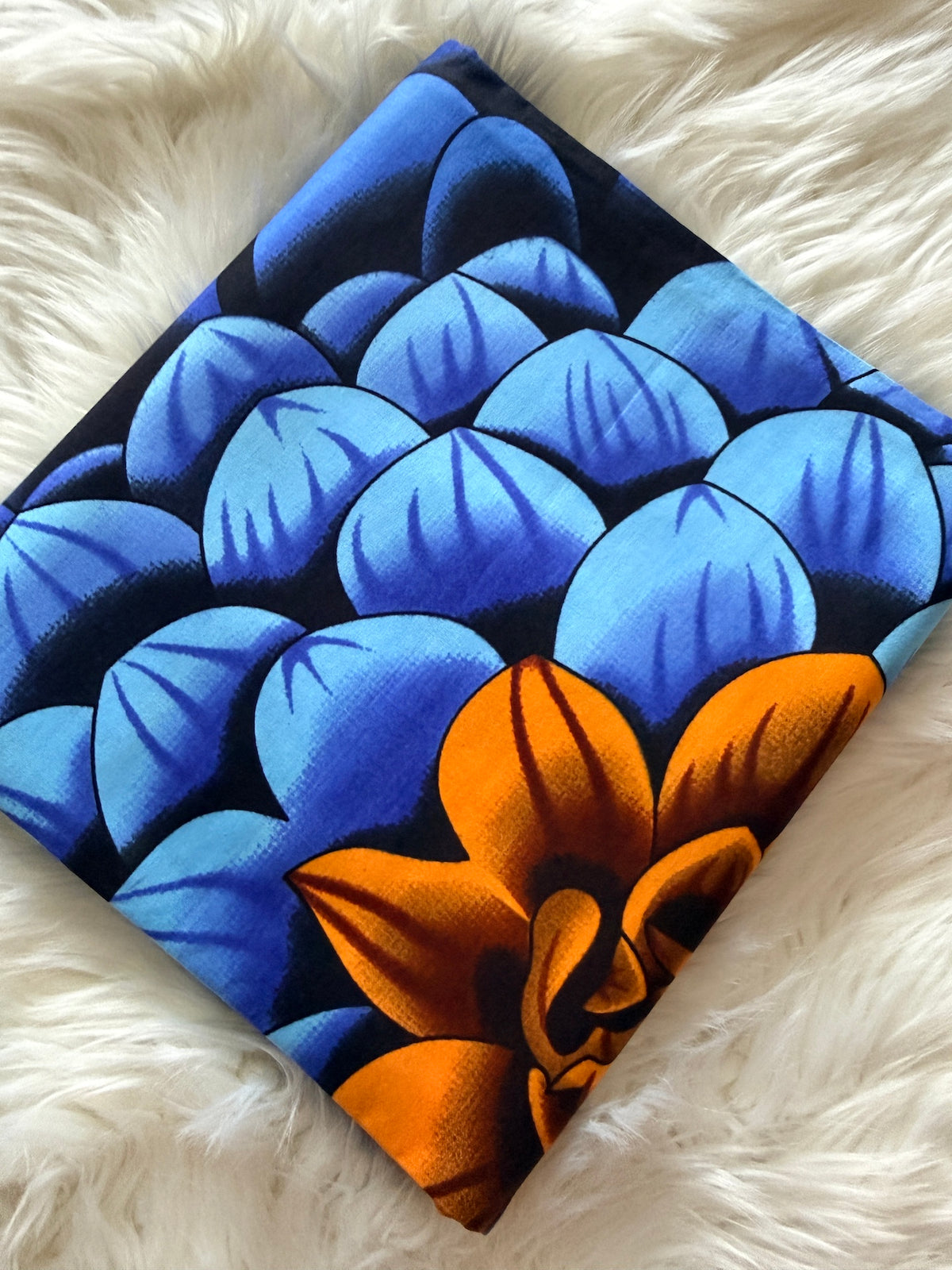 A square fabric item, likely a napkin or small scarf, featuring a bold pattern of blue and light blue scales with a large orange flower on a black background, displayed on a white faux fur surface.
