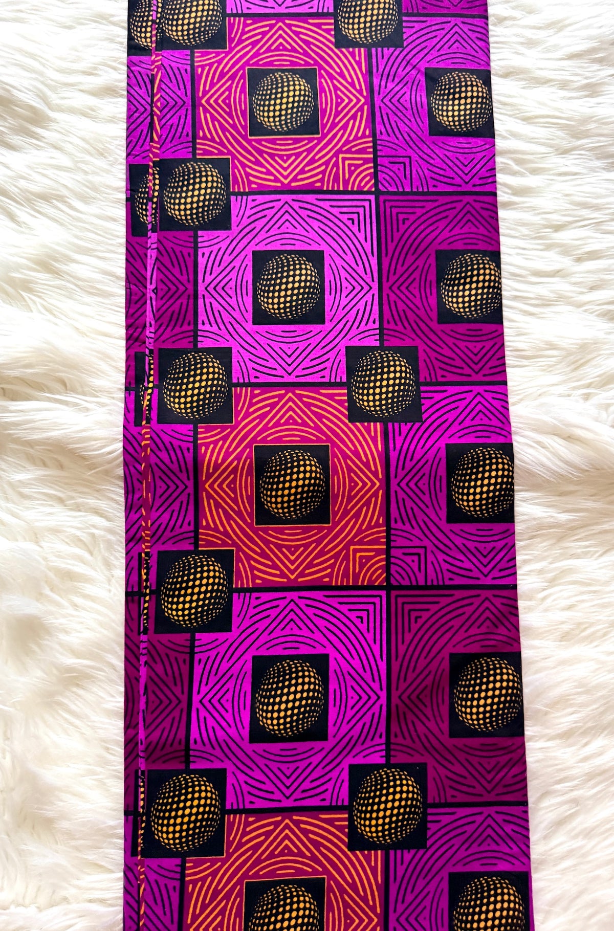 A long rectangular textile laid flat on a white faux-fur surface, featuring a bold geometric pattern in magenta, pink, orange, purple, black, and yellow with repeating cube motifs.