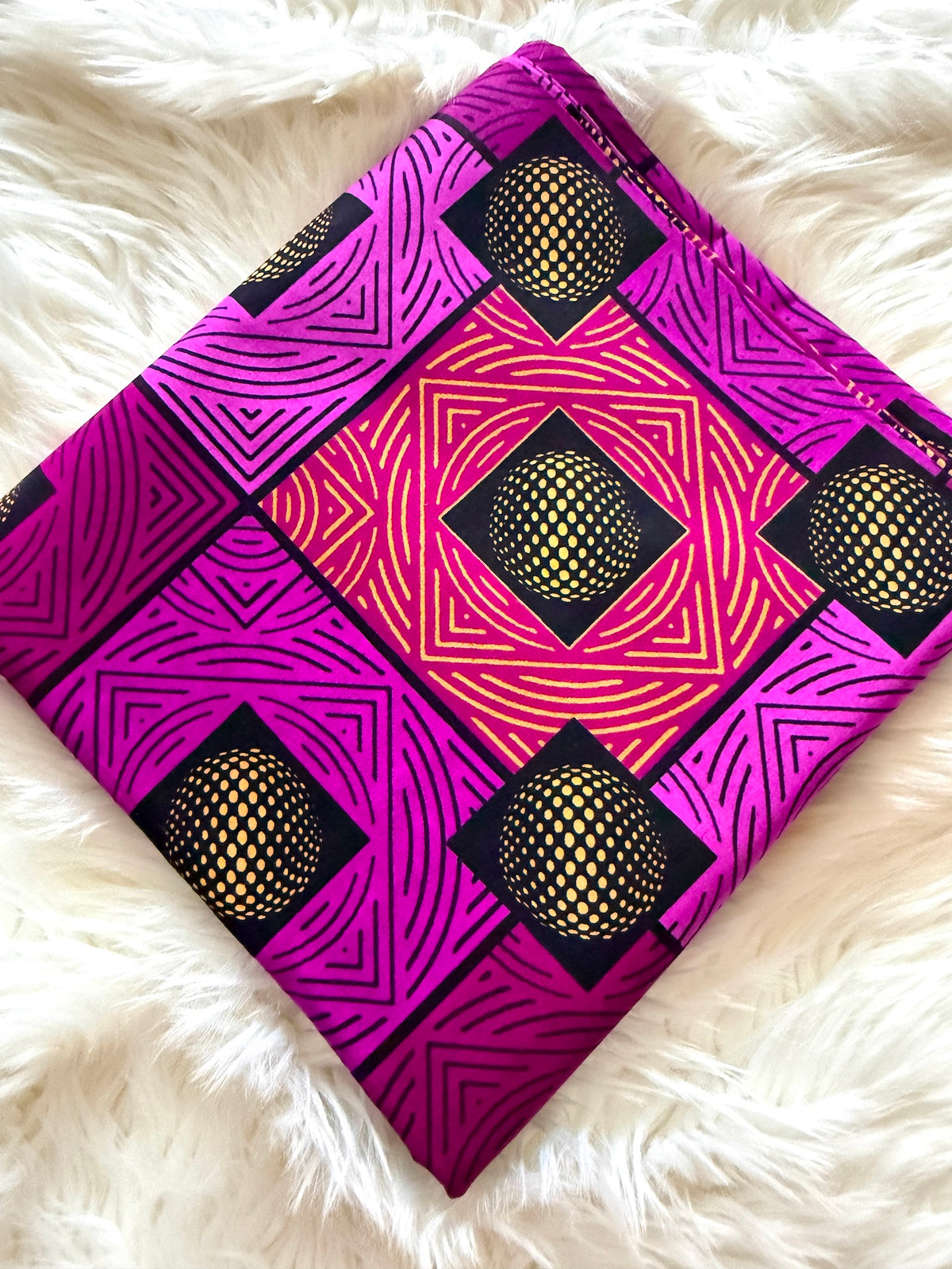 A square textile item, likely a scarf or bandana, featuring a vibrant magenta and pink African wax print (Ankara) pattern with black geometric lines and yellow-gold dotted sphere motifs, laid flat on a white faux fur background.