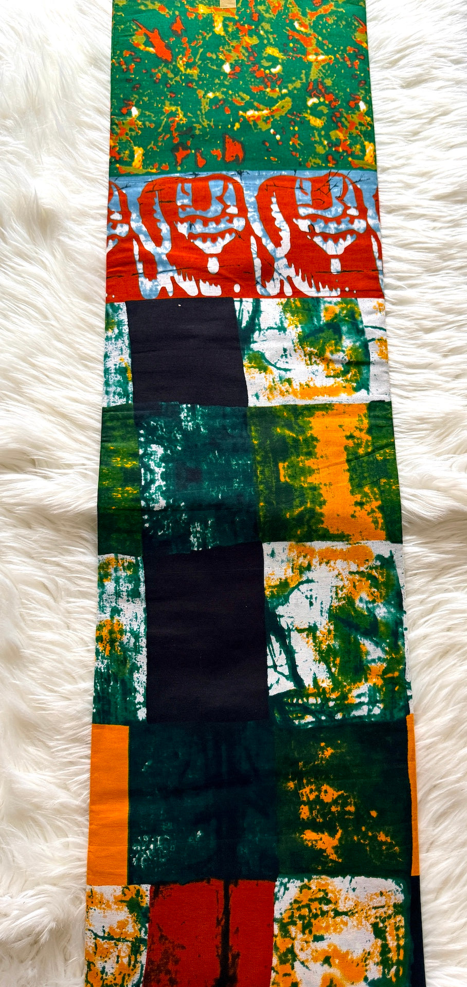 A long rectangular textile laid flat on a white faux-fur surface, featuring a patchwork-style print with green, orange, red, black, and white abstract and patterned blocks.