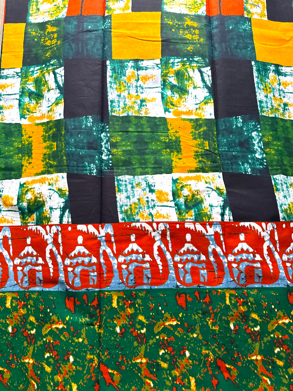 A close-up of a colorful patterned textile, likely a sari, featuring a checkered grid of green, yellow, black, and white squares, a horizontal red and white patterned band, and a green base with orange and yellow abstract floral prints at the bottom.