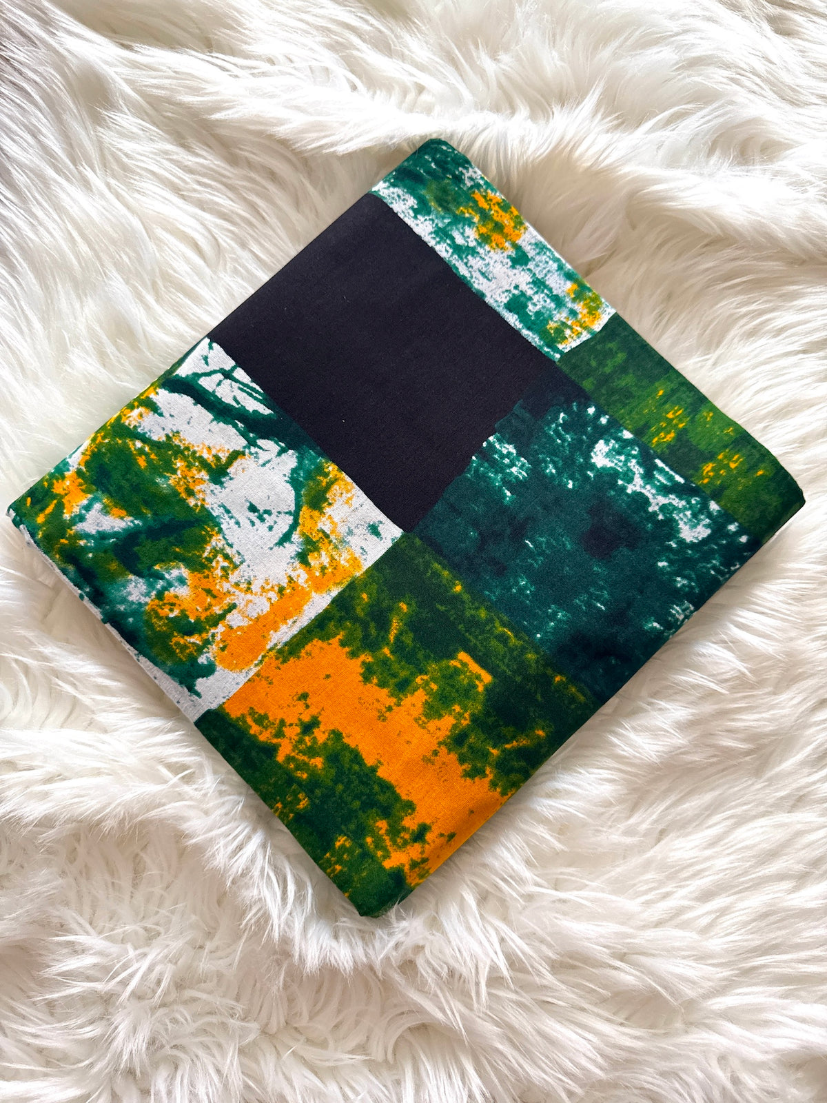 A square fabric item, likely a small blanket, pillow, or scarf, featuring an abstract patchwork print of green, orange, white, and black, laid flat on a white faux fur surface.