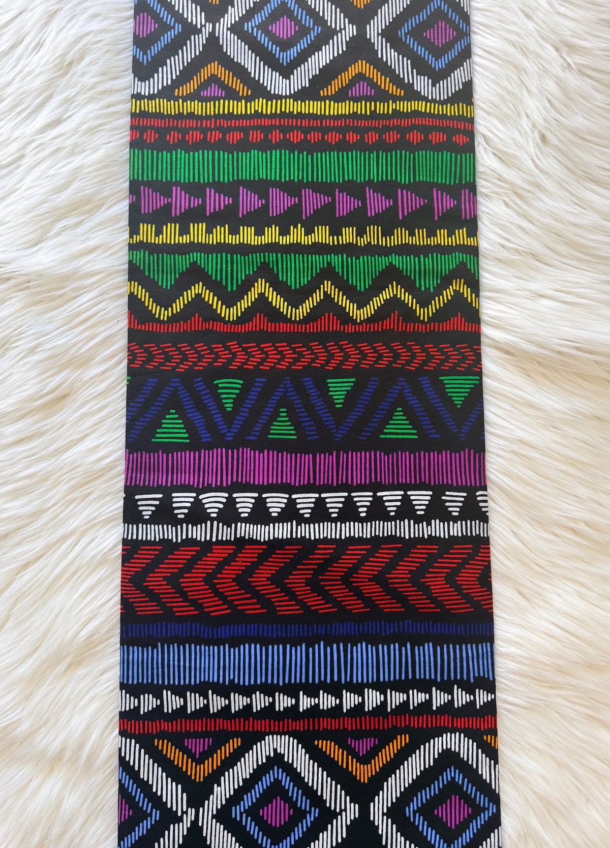 A long rectangular textile with a colorful geometric tribal pattern, laid flat on a white faux fur background.
