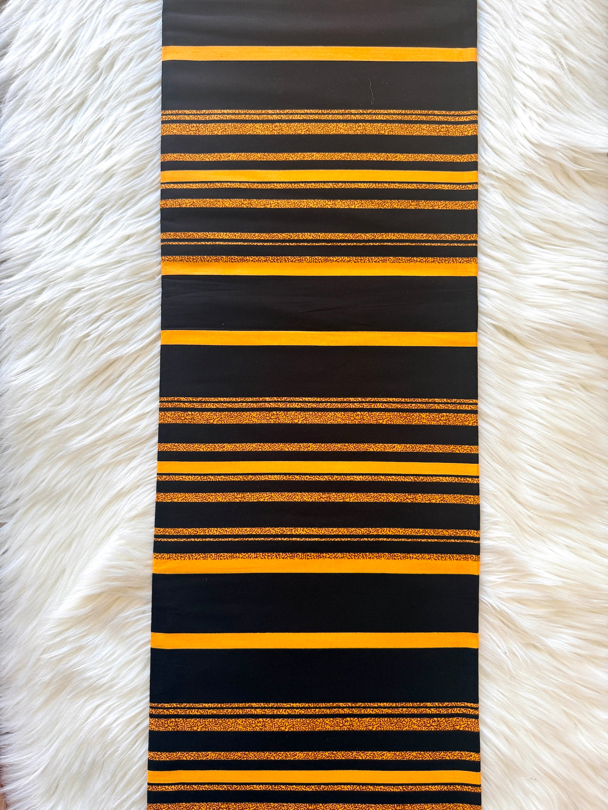 A long rectangular black table runner with horizontal stripes in bright yellow and shimmering gold glitter, laid flat on a white faux fur surface.