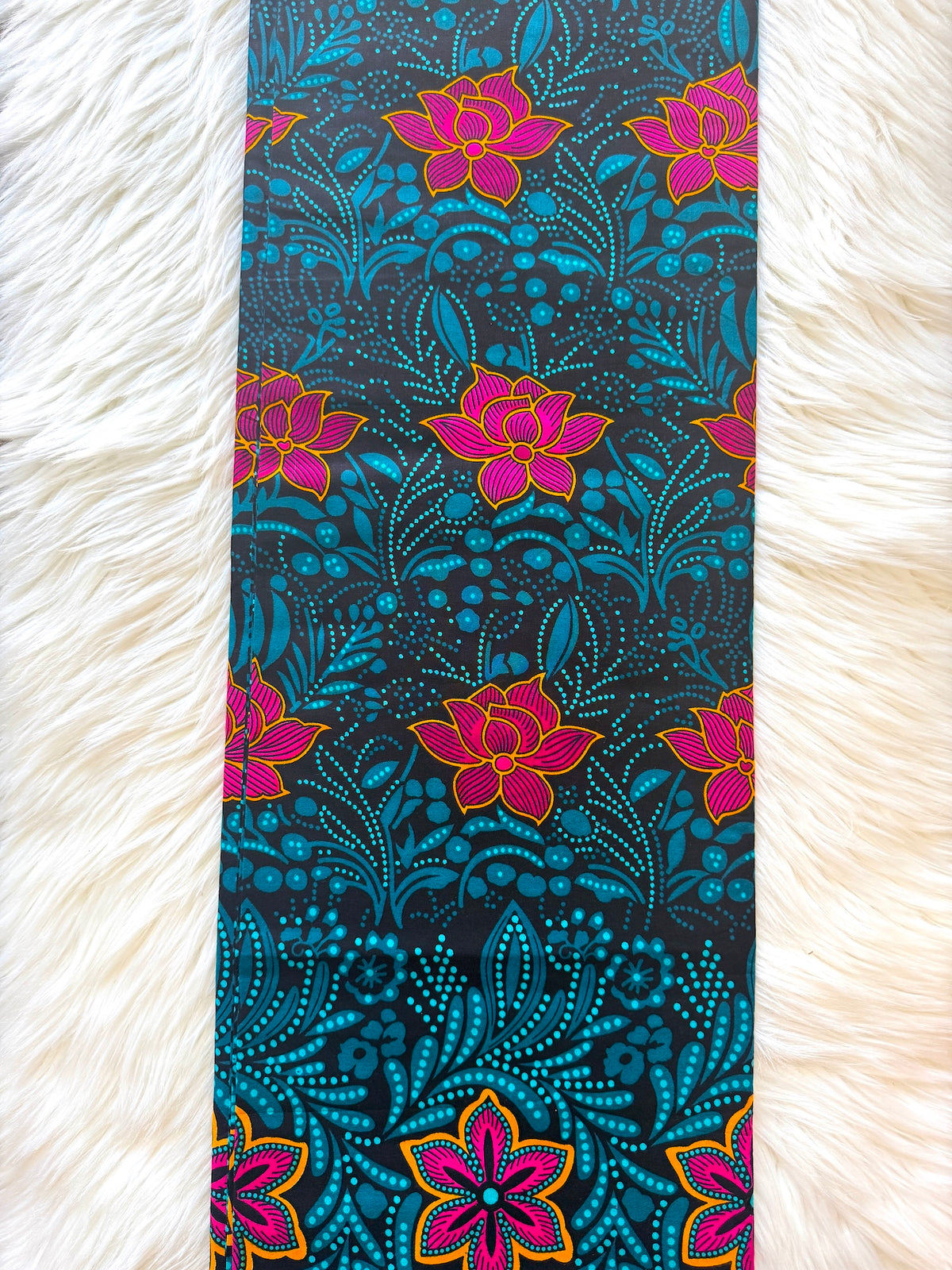 A long rectangular textile laid flat on a white faux-fur surface, featuring a bold floral and ornamental print in turquoise, black, magenta-pink, and yellow-orange.