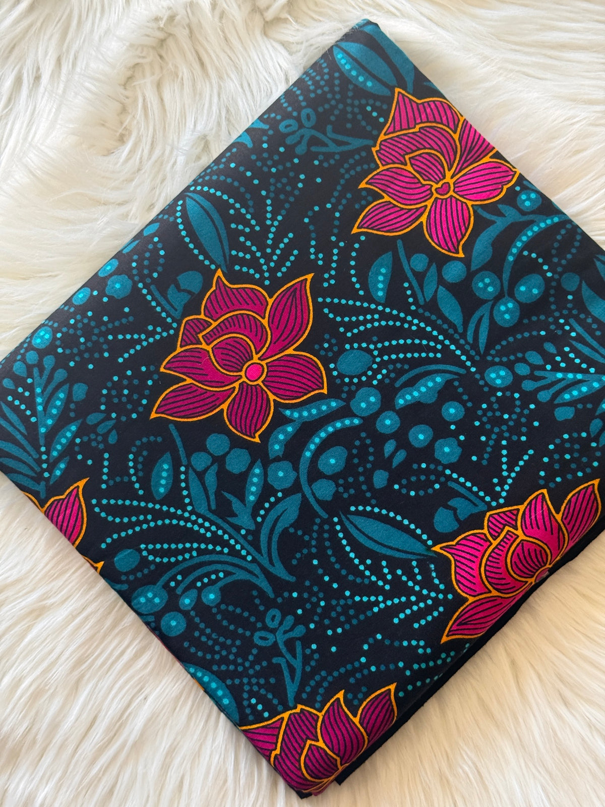 A square fabric item, likely a pillow cover or scarf, featuring a dark navy blue background with a vibrant pink and yellow lotus flower pattern and teal botanical dots, displayed on a white faux fur surface.