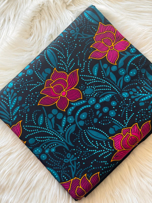 A square fabric item, likely a pillow cover or scarf, featuring a dark navy blue background with a vibrant pink and yellow lotus flower pattern and teal botanical dots, displayed on a white faux fur surface.