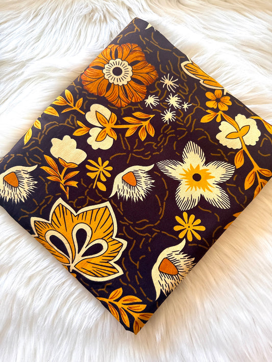 A square fabric or paper napkin with a retro floral pattern, laid flat on a white faux fur surface. The pattern features orange, yellow, and cream flowers and leaves on a dark brown background.