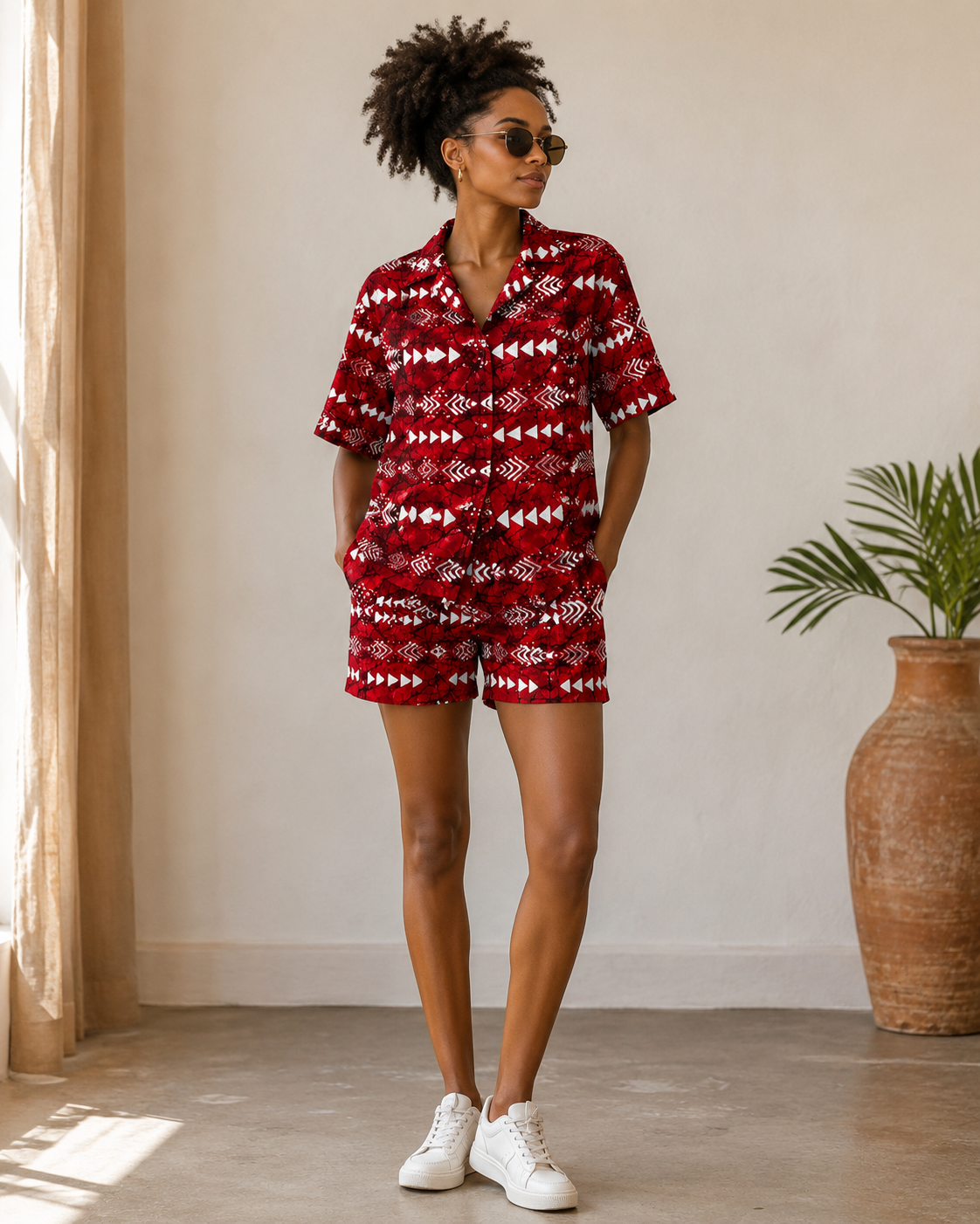 A woman modeling a matching red and white patterned short-sleeve shirt and shorts set in a bright indoor room with a large vase and plant in the background.