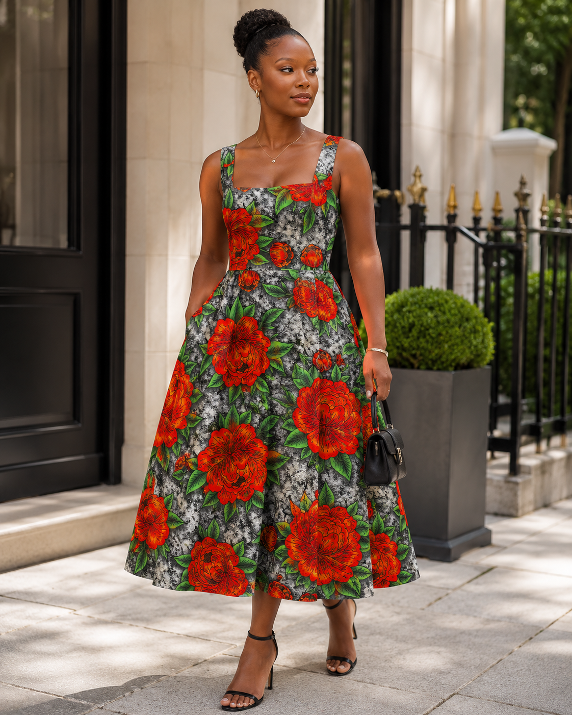 A woman stands on a sidewalk in front of a building entrance wearing a sleeveless midi dress with a bold floral print. The dress features large red flowers and green leaves on a dark background, paired with black strappy high-heeled sandals and a small black handbag.