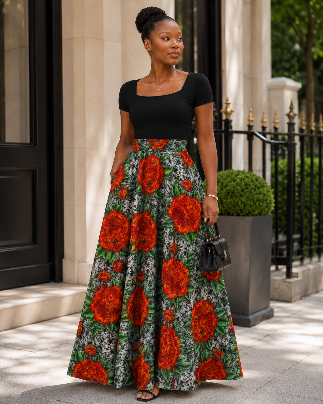 A woman stands outdoors on a sidewalk in front of a building entrance, wearing a black short-sleeve top and a full-length flared skirt with a bold red and orange floral print on a green background. She is holding a small black handbag.