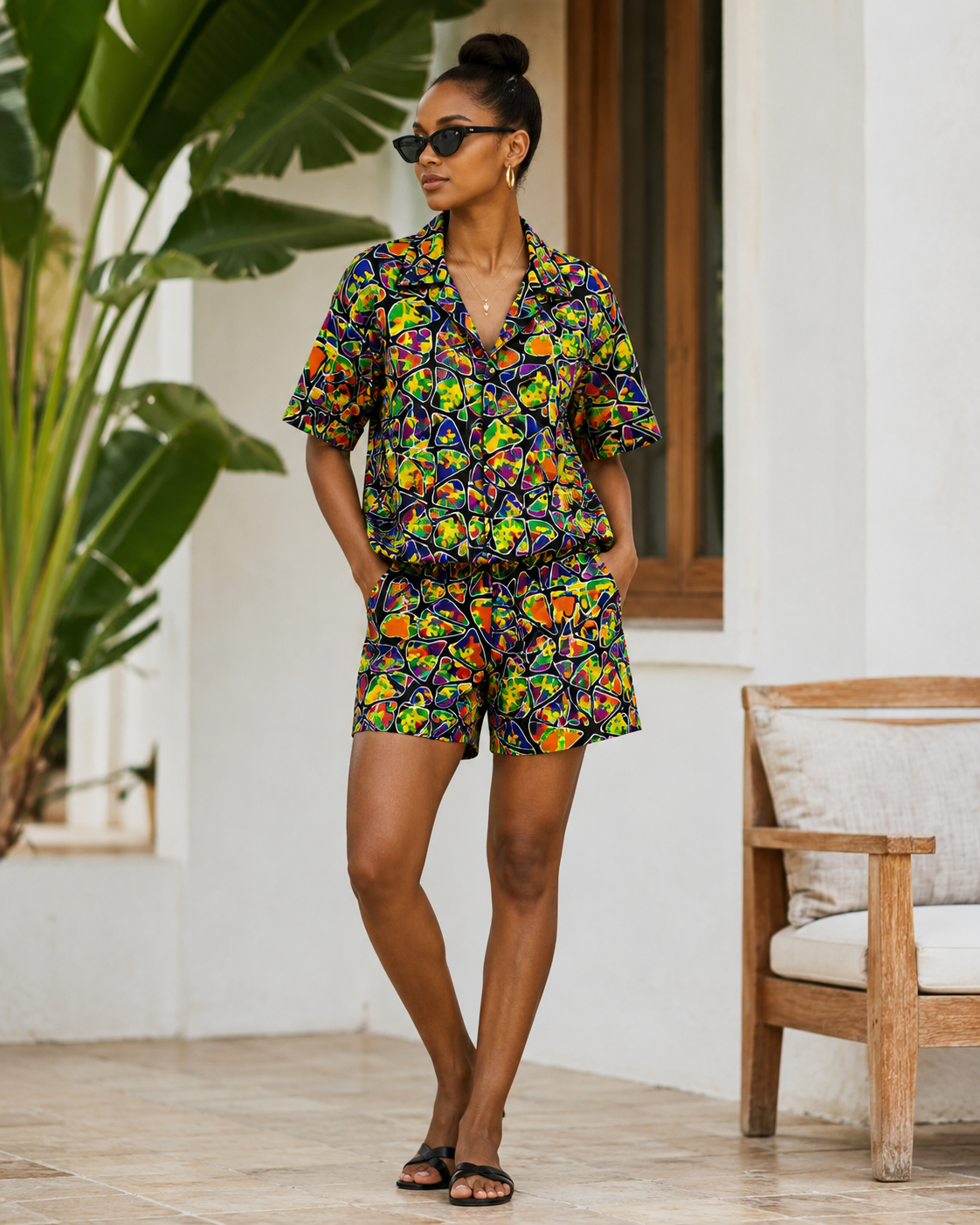 A woman modeling a matching two-piece short-sleeve shirt and shorts set with a vibrant multicolor floral print, set against a modern outdoor patio backdrop with a wooden chair and large green leaves.